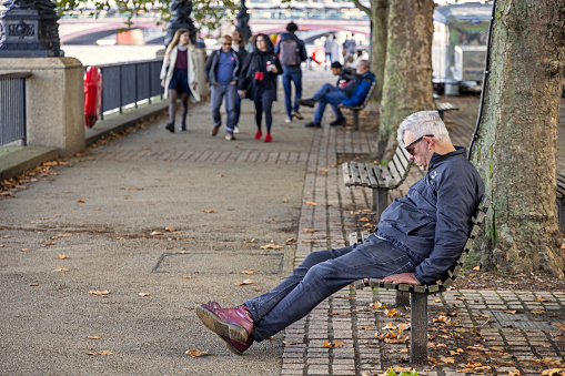 South bank of river Thames, London, England - October 27th 2023:  Middle aged man sleeping on a bench while the tourists walking at the riverside pedestrian area