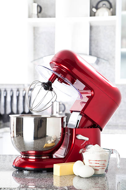 Red electric stand mixer on kitchen counter top A red electric stand mixer on kitchen counter top with eggs, butter and flour as ingredients for making a pie in the foreground. Focus in on the stand mixer while at the background can be seen a modern white kitchen out of focus.  electric mixer photos stock pictures, royalty-free photos & images