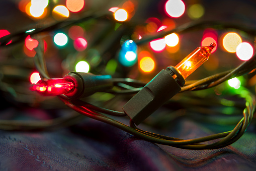 A group of Christmas lights on a string with a yellow light in focus in the foreground and the rest of the lights out of focus in the background.
