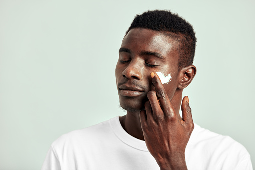 Black guy with youthful skin applying a moisturizing cream on his face to keep his skin soft and smooth. Handsome young African man maintaining his natural beauty with a skincare routine.