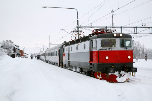 The temperature hovers well below zero as a passenger train stops at a snow-covered railway station in Swedenaas Arctic Circle.  Passengers gather near the train in the background.  The train provides some bright color in an all-white scene with the tracks and station all covered in fresh snow.  The train has snow and ice on its front and sides.  The headlights are lit.