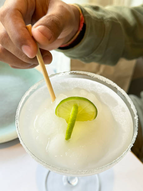 istock Close-up image of restaurant scene table with unrecognisable person using wooden swizzle stick to stir frozen Margarita tequila based cocktail from margarita glass with salted rim, lime slices garnish, elevated view, focus on foreground 1849737468