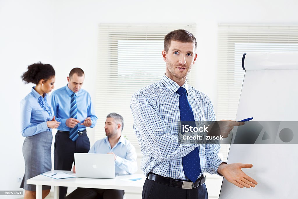Business meeting Focus on businessman giving presentation on the flip chart and looking at camera with his colleagues working in the background. Adult Stock Photo