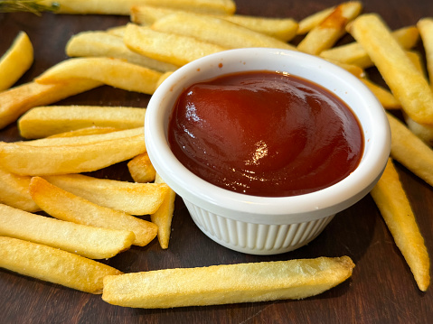 Stock photo showing close-up, elevated view of pile of crispy, golden, French fry chips and bowl ketchup of on wooden table.