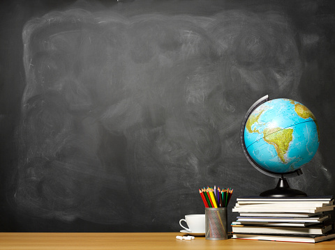 Globe of the world on a stack of books and pencils on a teacher's school desk with copy space on a blackboard