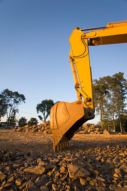 A sideview of the large bucket on a yellow backhoe / digger with the bucket in rocks. Photo taken during golden hour with clear blue sky.Lightbox of Building and Construction: