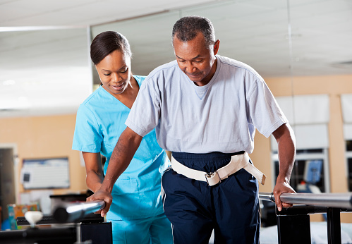 A female physical therapist assists an older male patient while doing gait training.  The man is holding on to railings, while the woman is behind him.