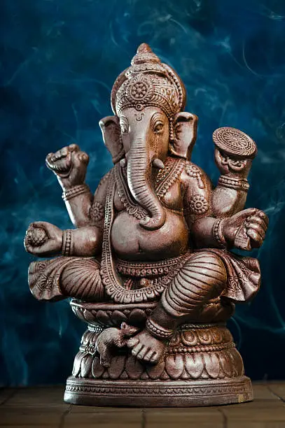 Deity of Ganesha from India on a blue background. Some incense are making smoke in the back and it looks like a shrine in a temple.