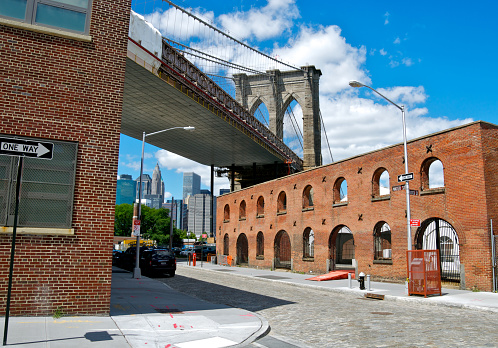 The Brooklyn Bridge seen from the perspective along Water Street in the DUMBO-Brooklyn Heights neighborhood of Brooklyn. In the distance portions of the skyline of Lower Manhattan can be seen on this sunny summer day in Brooklyn, New York City.