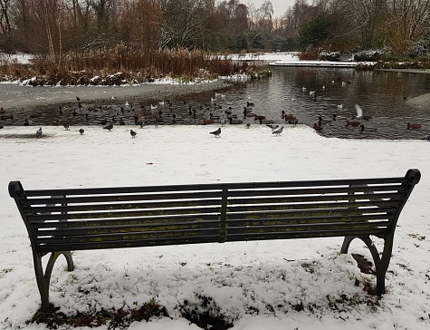 A metal seat park bench on a snow covered ground with a partly frozen pond view, Glasgow Scotland England UK
