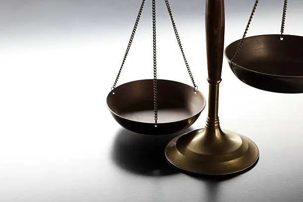 A justice scale on a simple gray background.  The scale is partially silhouetted as a strong backlight obscures some of the finer details.  Scale is place on far right hand side of image leaving ample negative space for copy. Image can be flipped horizontal to accommodate alternative composition needs.