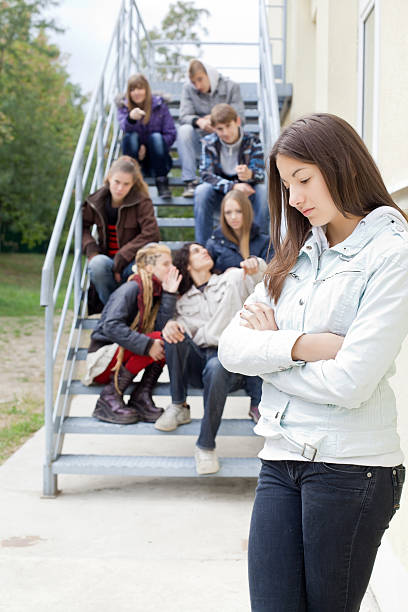 Unhappy teenage girl, her classmates gossiping on background. "Unhappy teenage girl, her classmates gossiping on background, pointing with fingers on her." slander stock pictures, royalty-free photos & images