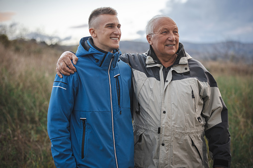 Teenage grandson and his grandfather walking together in nature, both wearing warm jackets, smiling and having a good time