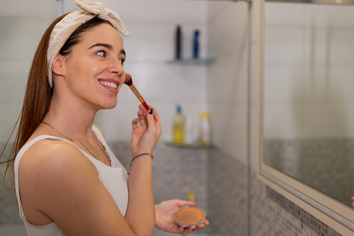 A side view of a young woman with a brush in her hand, applying blush on her cheeks while looking at the mirror