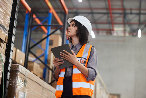 Young female engineer wearing a hardhat and reflective vest tracking shipments using a digital tablet while working in a shipping and distribution warehouse