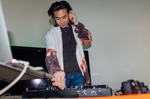 A young man wearing headphones on his head, using a laptop and mixing music on his DJ decks to supply music to a house party in North Shields, North East England. He is smiling while listening to the beat.