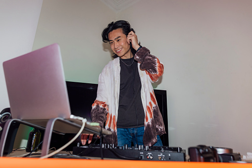 A young man wearing headphones on his head, using a laptop and mixing music on his DJ decks to supply music to a house party in North Shields, North East England. He is smiling while listening to the beat.