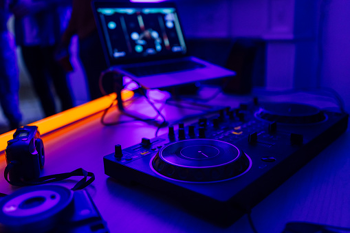A sound mixer on a table with a laptop in the background during a house party in North Shields, North East England. There are LED lights on making the whole room blue with an orange LED light on the table.