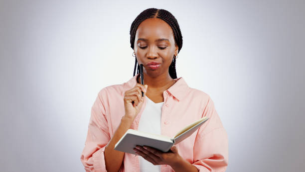 istock Woman, notebook and thinking of ideas, learning and studying with solution or goals on a white background. African student problem solving, reading notes and why face, confused or planning in studio 1849973341