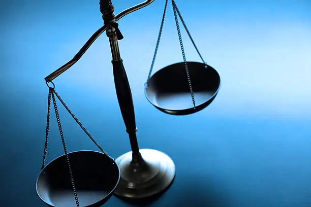 A justice scale on a simple blue background.  The scale is partially silhouetted as a strong backlight obscures some of the finer details.  Scale is place on left hand side of image leaving ample negative space for copy. Image can be flipped horizontal to accommodate alternative composition needs.
