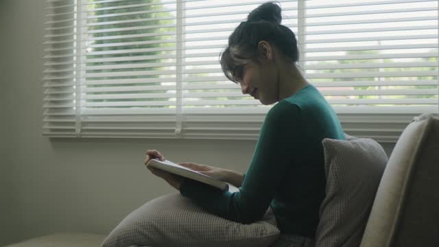 Young asian woman sitting on modern sofa in front of window relaxing in her living room reading book and drinking coffee or tea at home