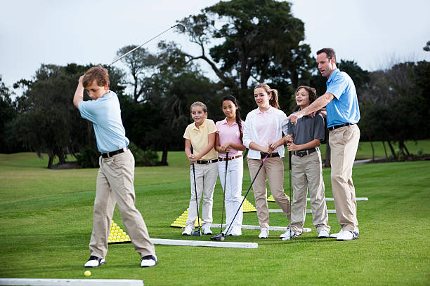 Golf pro teaching group of children (10 to 14 years), on practice range.  Focus on instructor and children watching.