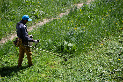 Tula, Russia - May 19, 2020: lawnmower man with string trimmer trimming grass at sunny day