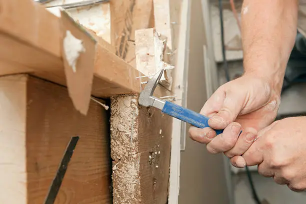 "A carpenter is using a nail puller in a stairwell at a house remodeling project. Text on the side of the tool reads, Warning: Always wear safety goggles. The workers hands are dry with worn fingernails and some cracks."