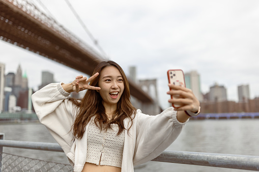 Two Chinese girlfriends taking a selfie for a memory. They are looking at the NYC Manhattan financial district skyline from the Brooklyn side