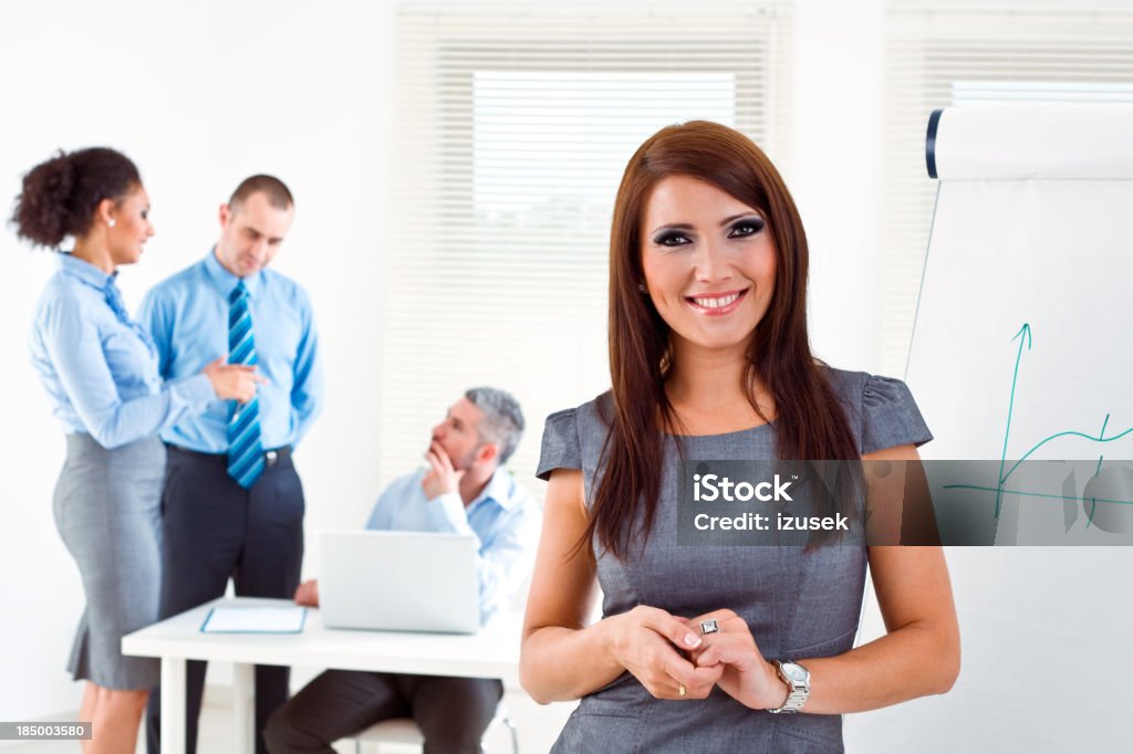Businesspeople at work Focus on the young businesswoman standing next to the flip chart and smiling at camera with her colleagues working in the background. Adult Stock Photo