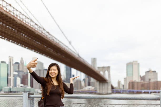 istock Chinese girl exploring NYC, taking selfie under the Brooklyn Bridge 1850036378