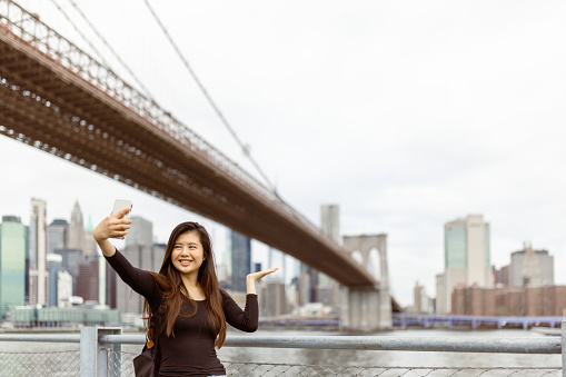 Two Chinese girlfriends taking a selfie for a memory. They are looking at the NYC Manhattan financial district skyline from the Brooklyn side