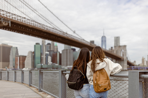 Two Asian girlfriends, students exploring Brooklyn Bridge, looking at the Lower Manhattan skyline from the bridge. They are on touristic trip to a NYC
