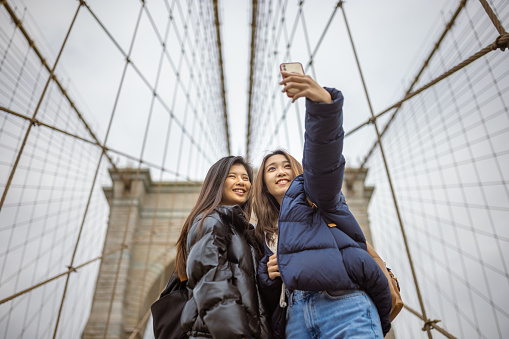 Two Asian girlfriends, students exploring Brooklyn Bridge, taking a selfie under the big, iconic pillar. They are on touristic trip to a NYC
