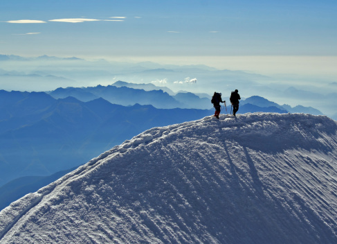 Two mountaineers on the narrow summit ridge of a mountain in the Swiss Alps. Chains of mountains are visible far below in the distance.