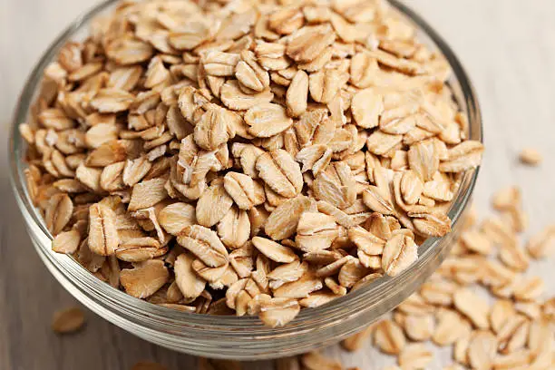 Close-up of a small glass bowl filled with rolled oats.  The oats are overflowing out of the bowl and have spilled onto the surface next to the bowl.  The bowl has rings around the edge, and the oats are raw and in a dry form.