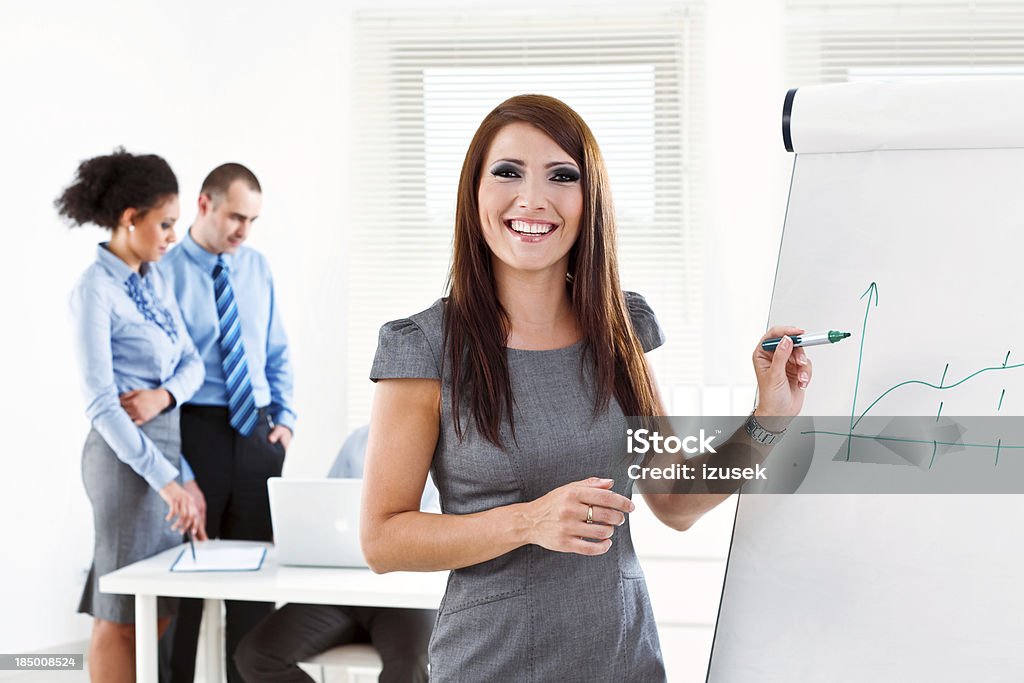Business presentation Focus on the young businesswoman giving presentation on the flip chart and smiling at camera with her colleagues working in the background. Adult Stock Photo