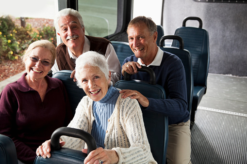 A group of four senior citizens riding in a van or shuttle bus, smiling at the camera.  The focus is on the woman with white hair in the foreground, wearing a white sweater.  Two women are sitting in front and two men are sitting directly behind them.  They are friends riding together, two couples.