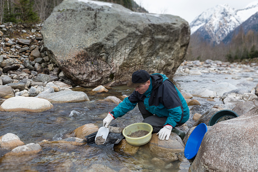 Gold panning in winter with the use of a sluice box, gold pan and sieve. Man is looking for gold in a mountain creek with a big boulder and blurry snow capped mountains as a background