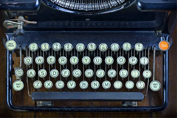 An antique manual black typewriter keyboard with its round keys and narrow oblong type hammers were common in offices before the invention of the electric typewriter. A bright orange "tab" key adds a bit of color to the keyboard's other keys of pale white.