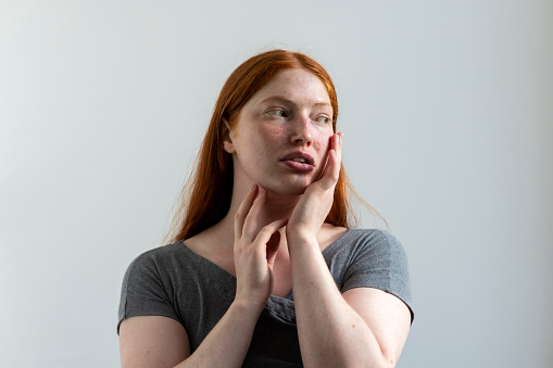 Close-up of a young female adult posing in the studio. She's standing faceing the camera but looking off to the side with her hands posed near her face.