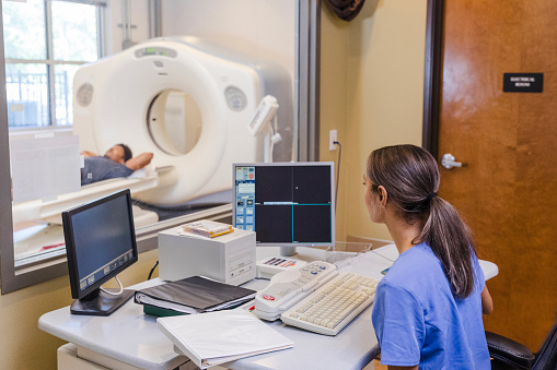 A young adult female radiographer instructs a young man who is preparing to have a CT scan. The radiograper talks to the man from the CT scan control room.