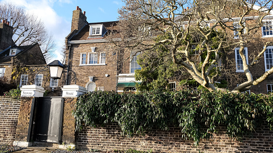 London, UK - 11.12.2023. Magnolia tree near the house by Thames riverside. Brick fence, stones. Fence wooden door. House behind the fence. Georgian medieval brick house. Residential house. Winter in London