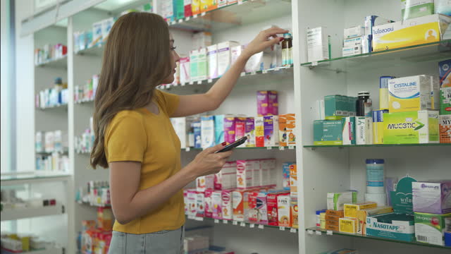 Young Woman Using Smart Phone Searching Medicine At Pharmacy