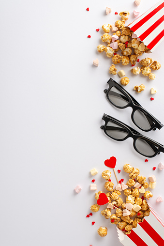 Valentine's Day cinema joy. Vertical top view of couple of 3D glasses, striped popcorn boxes, heart decor, marshmallow, and sprinkles on white background. Perfect for text or advertising