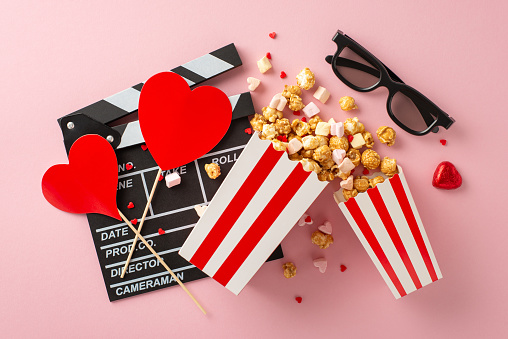 Cinematic love story on Valentine's. Top view picture showcasing clapperboard, 3D glasses, striped popcorn containers, hearts on sticks, candies, marshmallows, sprinkles on dreamy soft pink surface