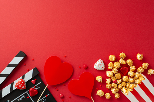 Hearts on Screen: Top view of a clapperboard, popcorn box, chocolates, sprinkles and heart decor, creating an enchanting atmosphere for the premiere of a romantic film on a red background