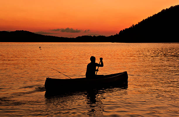 Canoe Lake Silhouette of canoe and man paddling and fishing at sunset on Mary Lake, Ontario at an orange glow sunset.  Muskoka sunset. huntsville ontario stock pictures, royalty-free photos & images