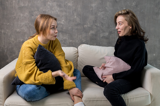 two young women are talking sitting on the couch. one woman is very surprised