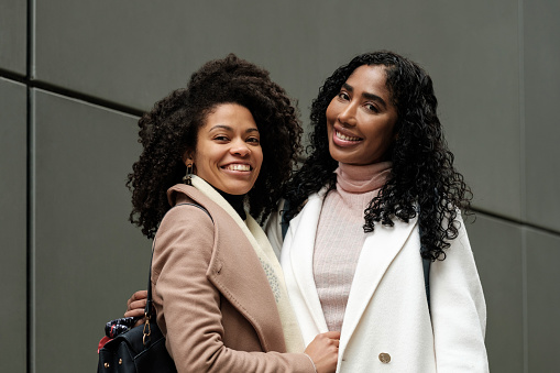 Carefree portrait of female friends posing in front of grey wall. It is winter and they are wearing thick coats. Their hairs are curly and they are smiling and looking happy.